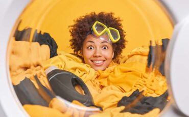 Happy pleased housewife smiles broadly enjoys washing loads washer with laundry uses liquid powder wears snorkeling glasses on forehead busy doing housework. Domestic responsibilities concept