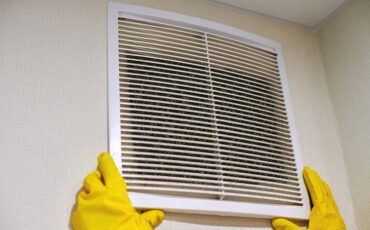 Hands in protective rubber gloves opening clogged air ventilation grill of HVAC with dusty filter to clean or replace it. Cleaning service concept.