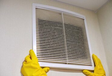 Hands in protective rubber gloves opening clogged air ventilation grill of HVAC with dusty filter to clean or replace it. Cleaning service concept.