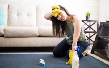 Young woman rubbing head wearing casuals and yellow gloves cleaning carpet with liquid soap